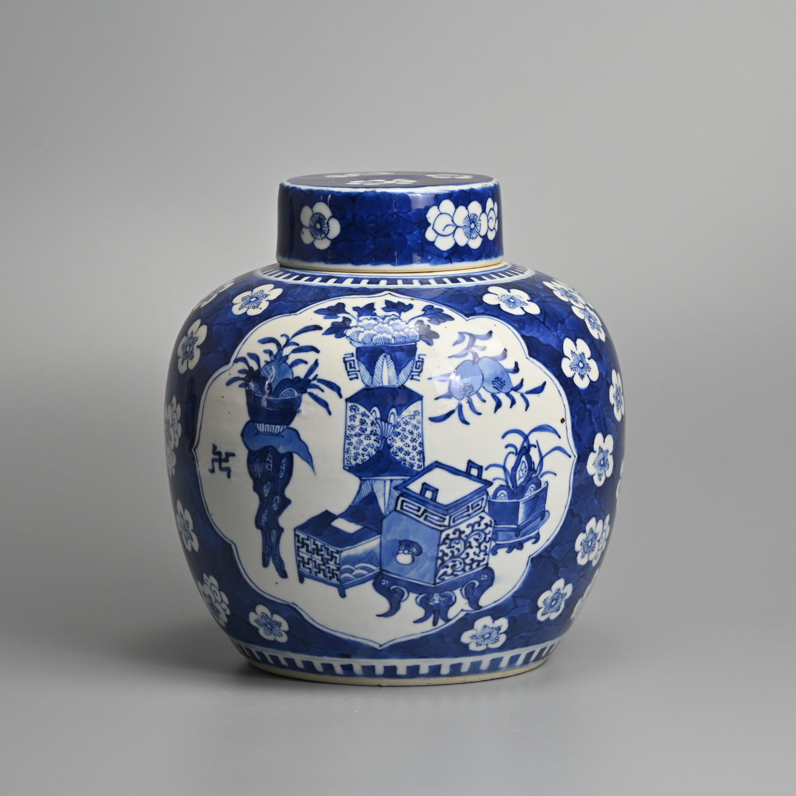 A LARGE CHINESE PORCELAIN BLUE AND WHITE GINGER JAR AND COVER, 19TH / EARLY 20TH CENTURY.: Painted in the Kangxi style with vases of flowers and fruit flanked by Buddhist swastikas within lappet-shaped panels, with prunus on a mottled blue ground, with a cork stopper, with double concentric