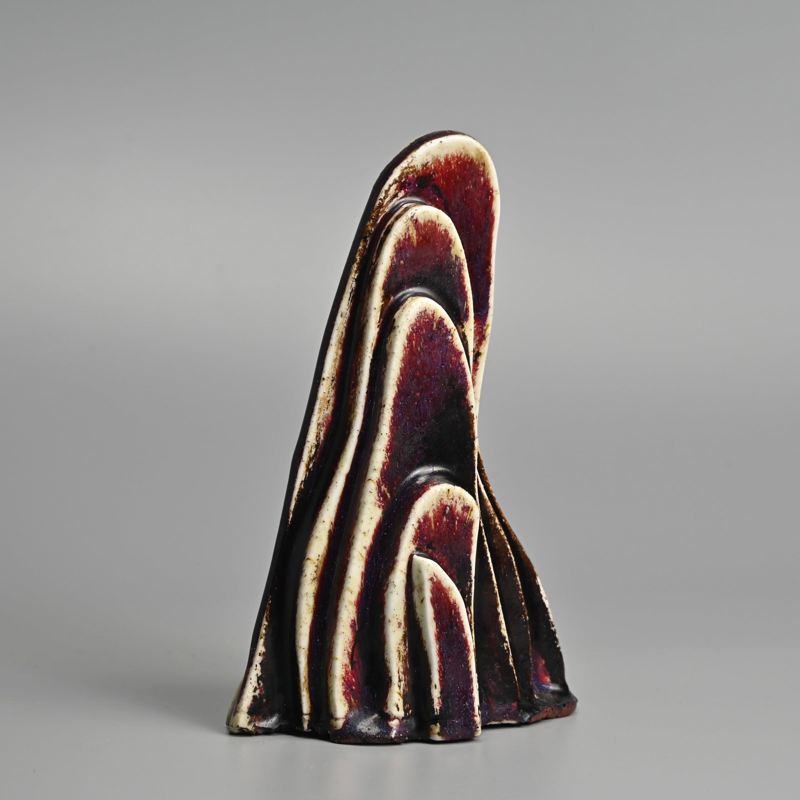 A CHINESE FLAMBE GLAZED PORCELAIN 'SCHOLAR'S DESK MOUNTAIN', QING DYNASTY.: In the form of a stone formation covered in a reddish glaze with purple streaks in places thinning to a off white colour to the edges. 19.5cm tall.