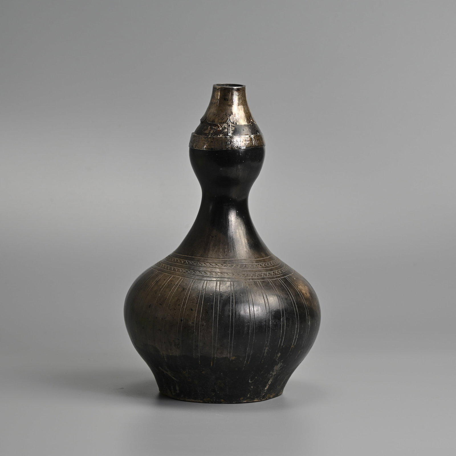 A MALAY DOUBLE GOURD FORM WATER FLASK, LABU, 19/20TH CENTURY.: Earthenware body covered in a thin charcoal coloured glaze decorated with incised lines and 'x' pattern around the lower half of the body. Mounted with a white metal section at the rim which has a ban