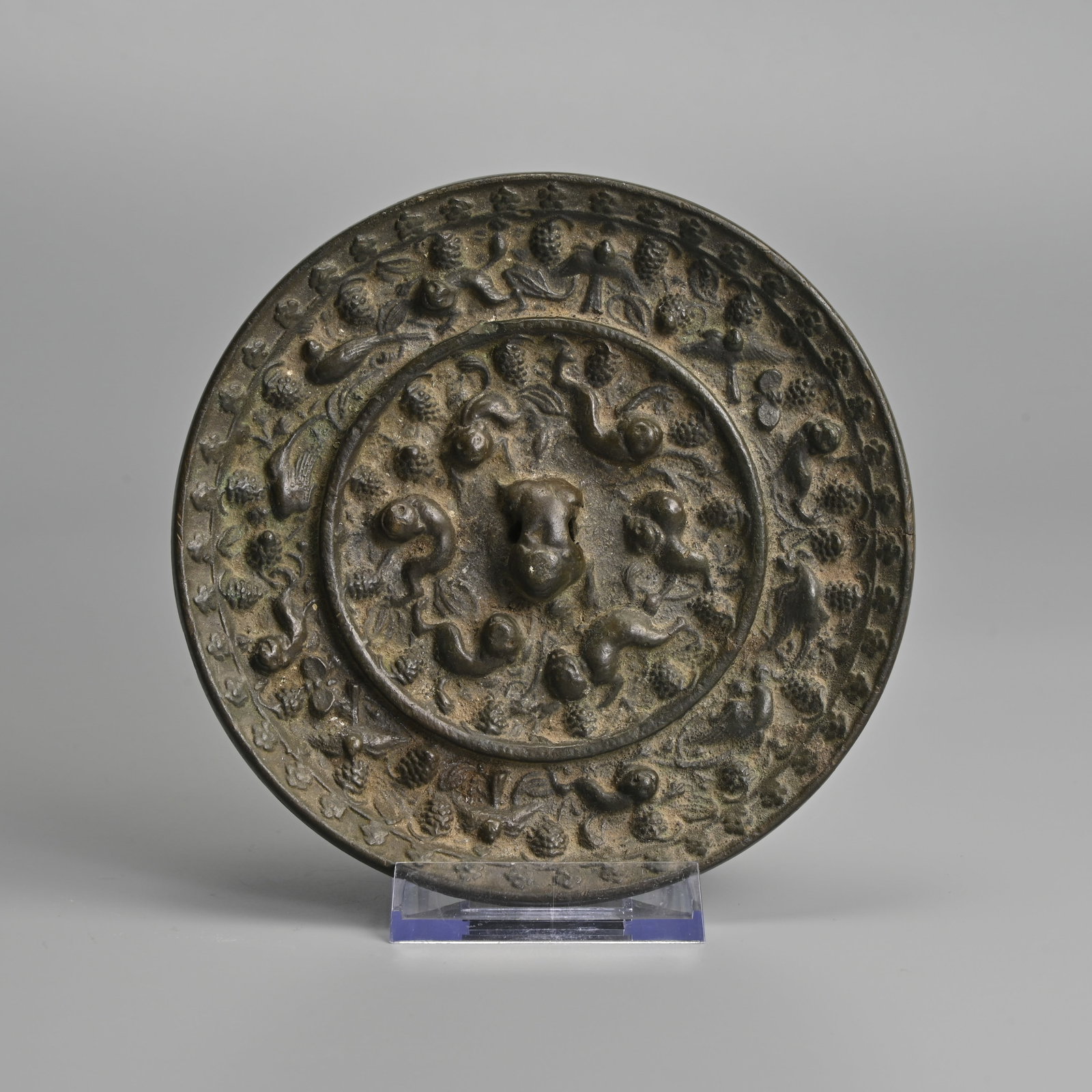 A CHINESE BRONZE 'LION AND GRAPEVINE' MIRROR, TANG DYNASTY (618-907 AD).: Cast with a central recumbent mythical beast knop, surrounded by a band of four recumbent lions alternating with fruiting grape vines. The outer border with a band of four lions, birds and grape vines