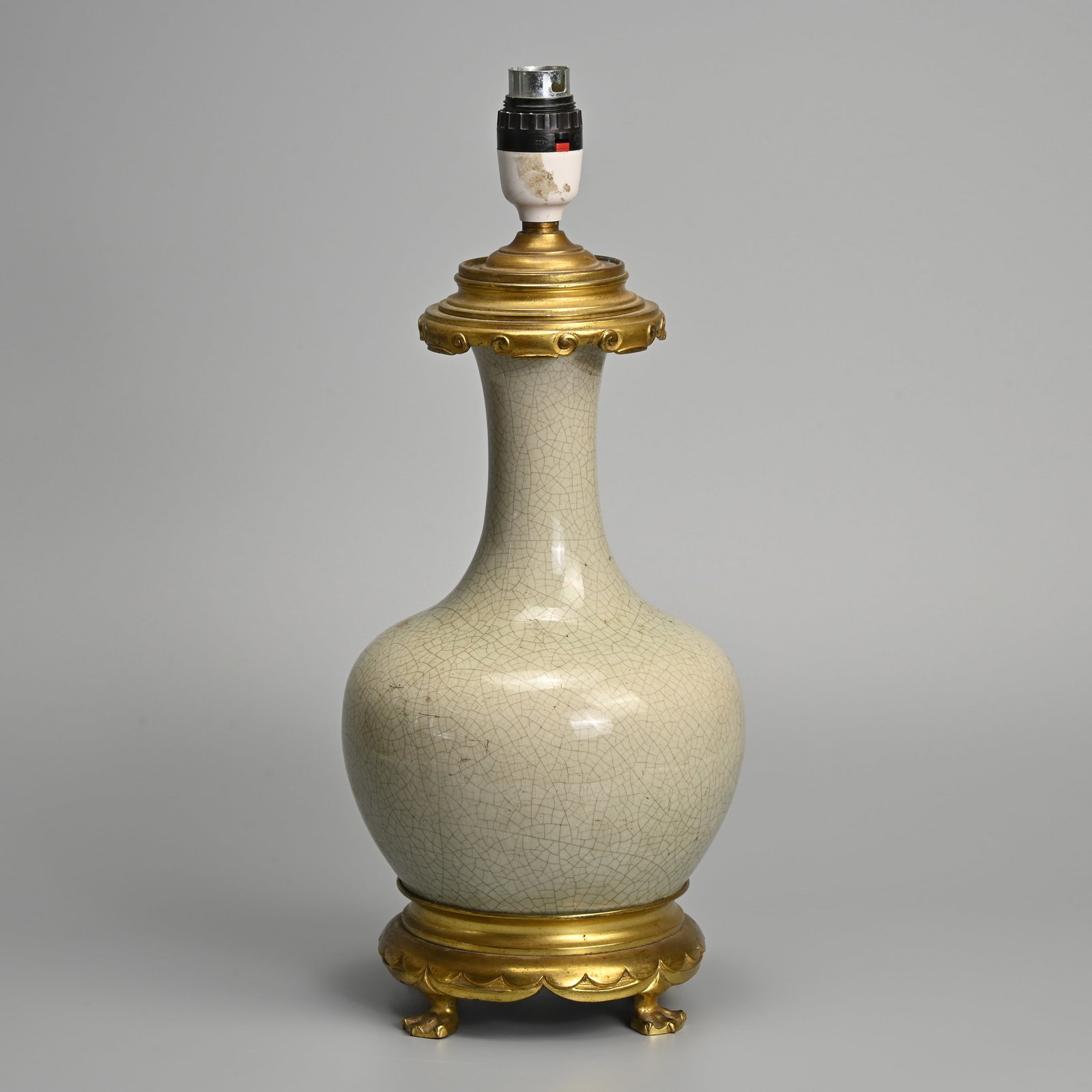 A CHINESE CRACKLE GLAZED PORCELAIN BOTTLE VASE CONVERTED INTO A LAMP.: A CHINESE CRACKLE GLAZED PORCELAIN BOTTLE VASE CONVERTED INTO A LAMP. The vase covered in a greyish crackle glaze mounted on a gilt metal base with four webbed feet. 38cm tall including lamp fitting.