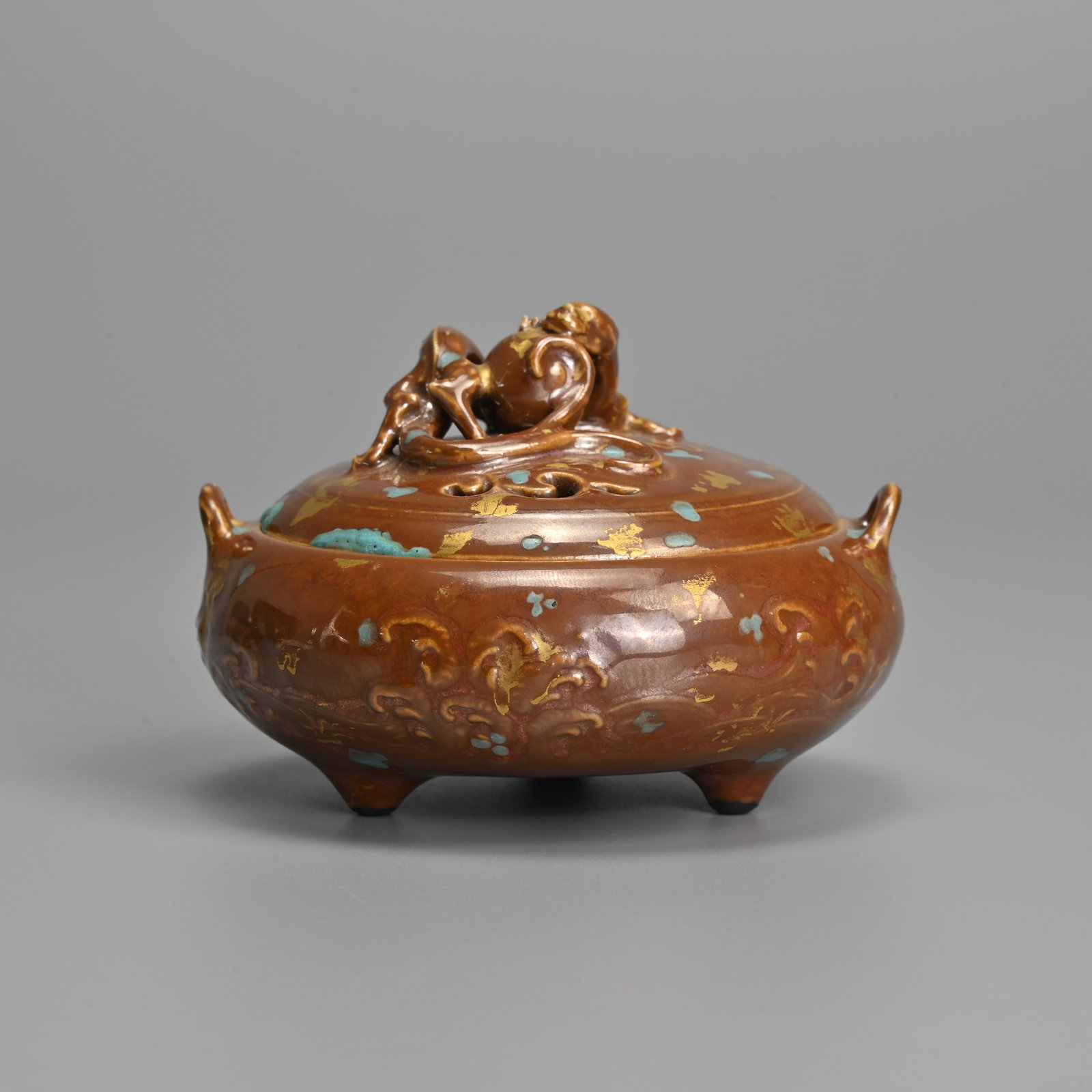 A SMALL CHINESE GILT AND TURQUOISE SPLASHED BROWN GLAZED PORCELAIN TRIPOD CENSER, QIANLONG MARK,: A SMALL CHINESE GILT AND TURQUOISE SPLASHED BROWN GLAZED PORCELAIN TRIPOD CENSER, QIANLONG MARK, 20TH CENTURY. Or squat form with two looped handles, moulded leafy scrolls to the exterior with a pierc