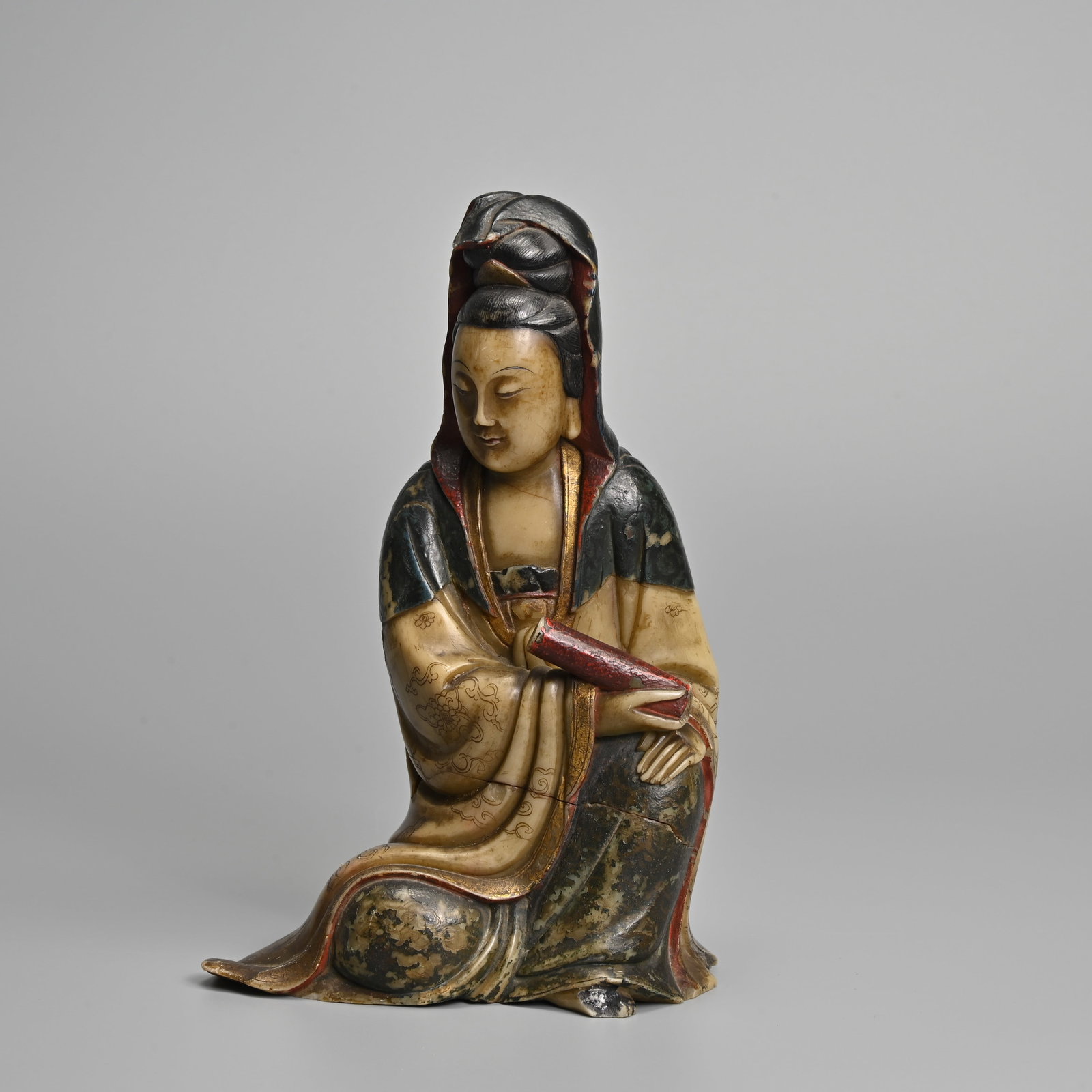 A CHINESE CARVED PAINTED SOAPSTONE FIGURE OF SEATED GUANYIN, 19TH CENTURY.: A CHINESE CARVED PAINTED SOAPSTONE FIGURE OF SEATED GUANYIN, 19TH CENTURY. The Goddess of Mercy seated dressed in robes incised with cloud motifs and her hear covered by a shawl, holding a scroll. Col