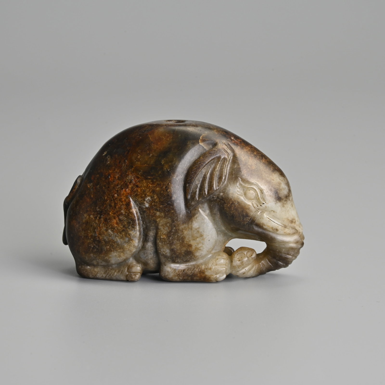 A CHINESE CARVED GREENISH GREY JADE RECUMBENT ELEPHANT PENDANT, QING DYNASTY.: A CHINESE CARVED GREENISH GREY JADE RECUMBENT ELEPHANT PENDANT, QING DYNASTY. The elephant front facing with its legs tucked under its body. A single aperture through the centre. 7.6cm length x 5cm ta