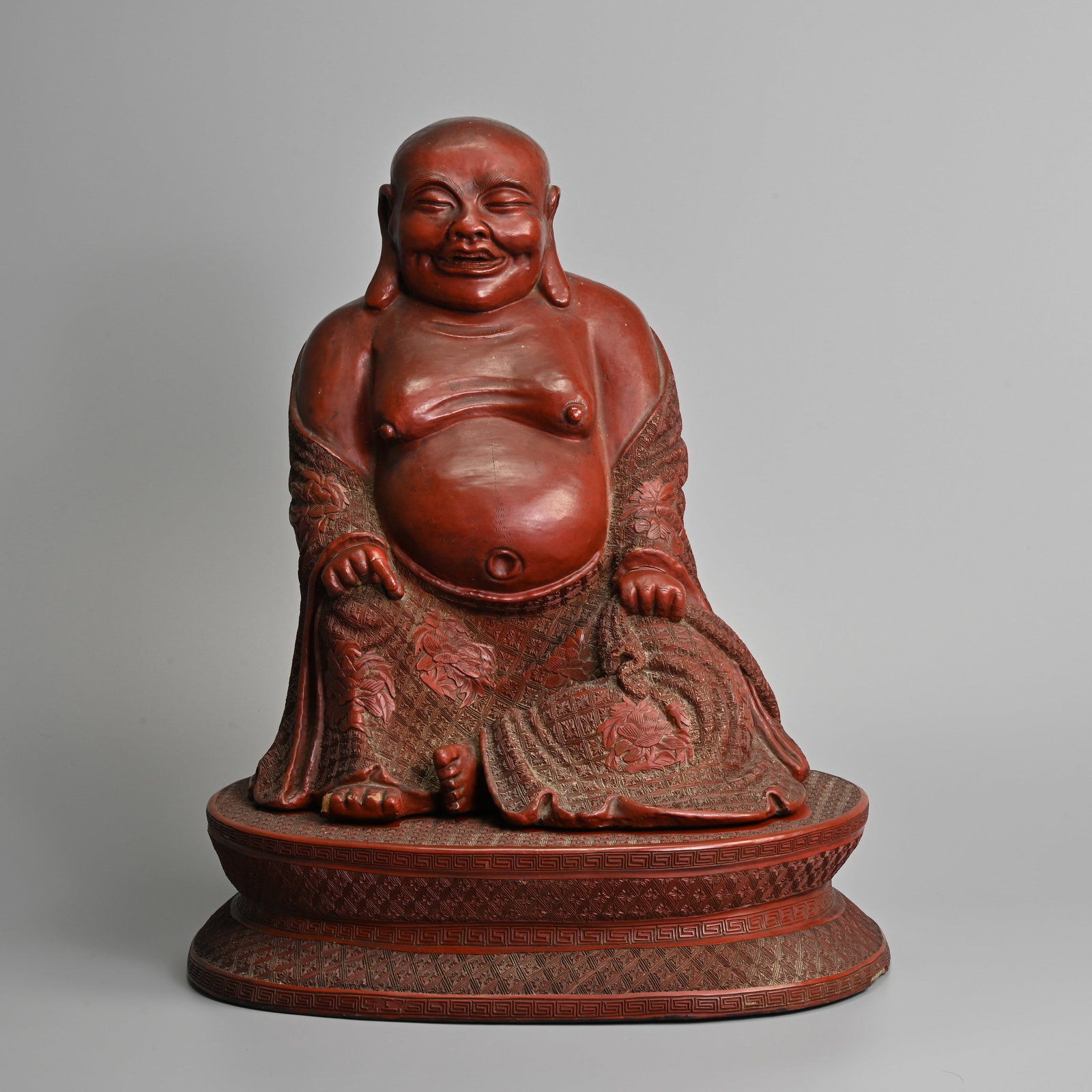 A LARGE CHINESE RED LACQUER FIGURE OF BUDAI ON SEPARATE STAND, LATE QING DYNASTY.: A LARGE CHINESE RED LACQUER FIGURE OF BUDAI ON SEPARATE STAND, LATE QING DYNASTY. The laughing Buddha seated wearing a loose robe decorated with floral patterns, open to reveal his chest and stomach. 