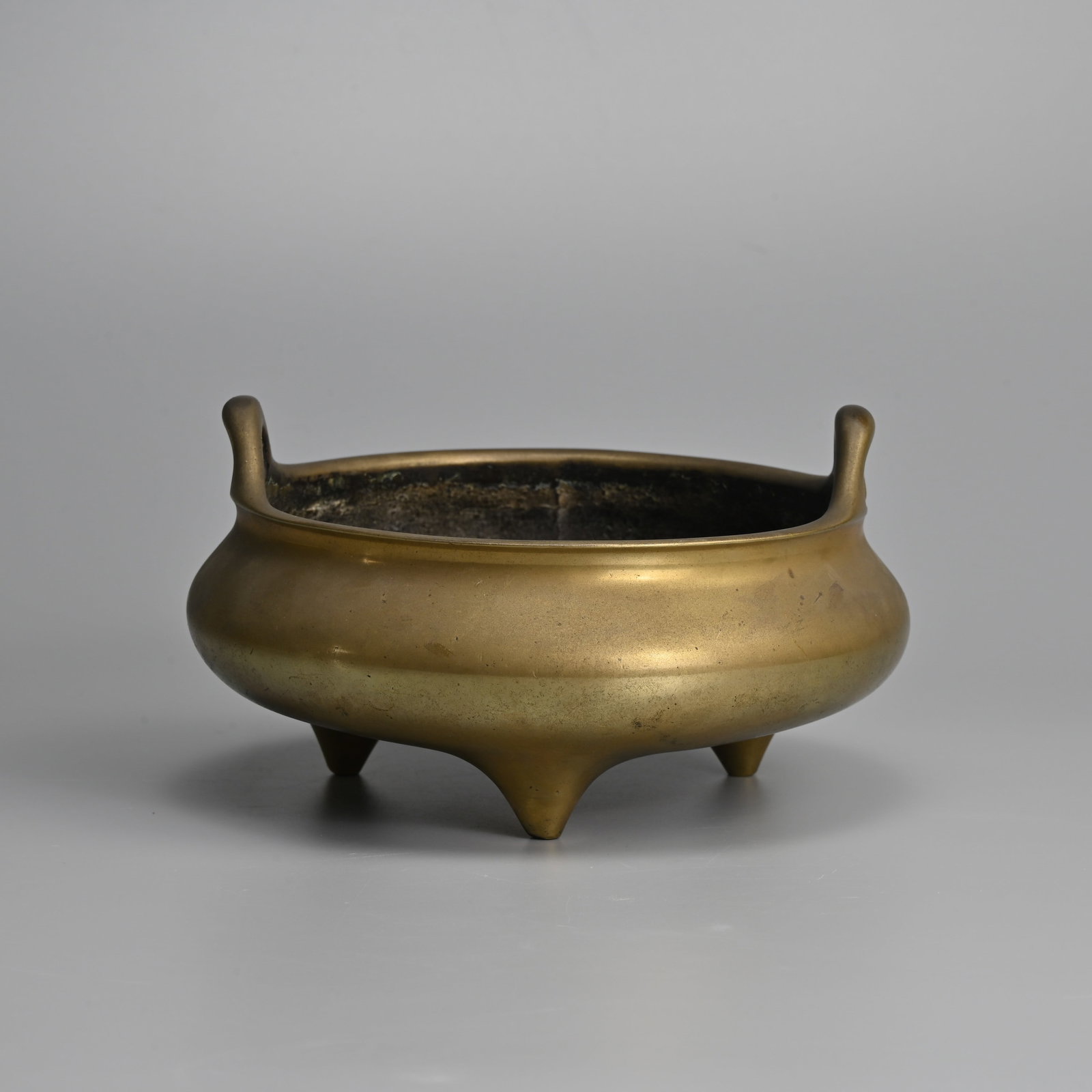 A CHINESE POLISHED BRONZE TRIPOD CENSER, XUANDE MARK, QING DYNASTY.: A CHINESE POLISHED BRONZE TRIPOD CENSER, XUANDE MARK, QING DYNASTY. Raised on three coned feet with looped handles. Relief six character mark of Xuande to base. 18.5cm diam. overall. Weight 2260 grams