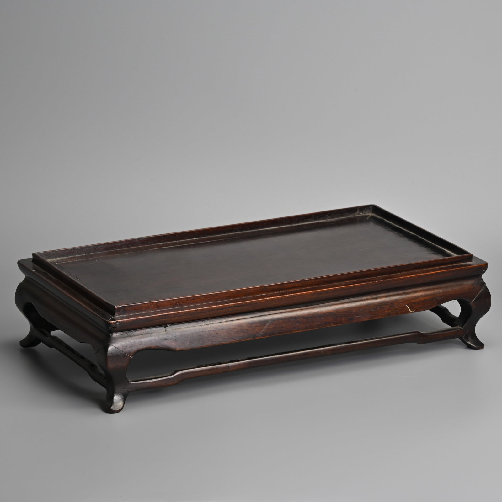 A CHINESE HARDWOOD TABLE STAND, EARLY 20TH CENTURY.: A CHINESE HARDWOOD TABLE STAND, EARLY 20TH CENTURY. Of rectangular form raised on four cabriole legs with curved frieze and stepped stretchers. The top fitted with a raised border. 41cm length x 22cm 