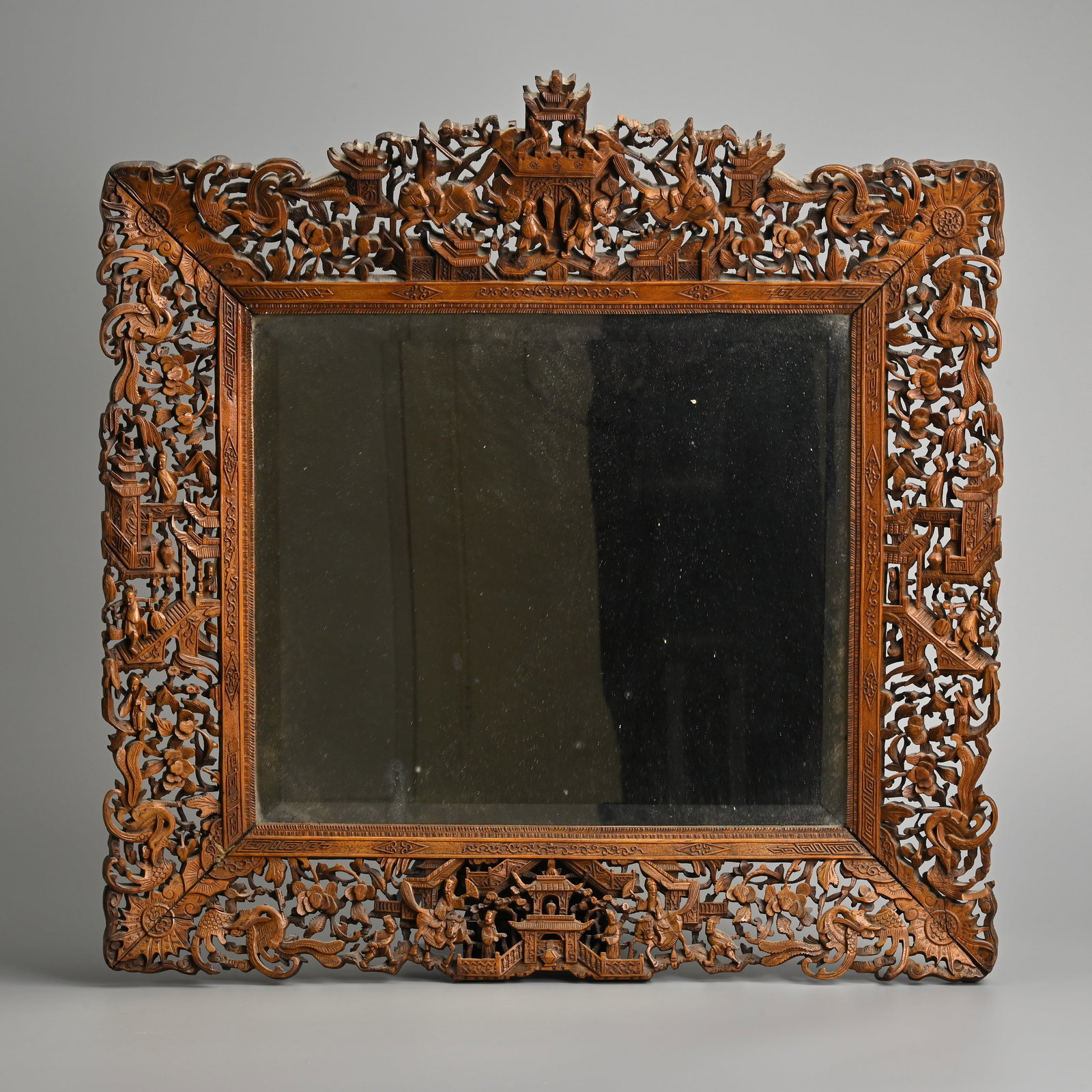A CHINESE CARVED WOOD FRAMED MIRROR, CIRCA 1900.: A CHINESE CARVED WOOD FRAMED MIRROR, CIRCA 1900. The pierced frame intricately carved with various figures on buildings with phoenix and floral scrolls. 56.5cm wide x 57cm tall.