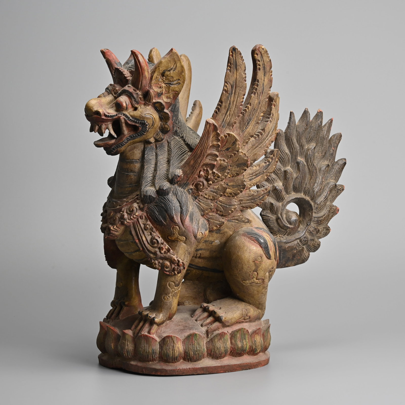 A LARGE BALINESE CARVED WOOD FIGURE OF A WINGED LION, SINGA BERSAYAP, INDONESIA, EARLY 20TH CENTURY.: A LARGE BALINESE CARVED WOOD FIGURE OF A WINGED LION, SINGA BERSAYAP, INDONESIA, EARLY 20TH CENTURY. The mythical guardian lion seated on a lotus base, polychrome decorated with a separately carved ta