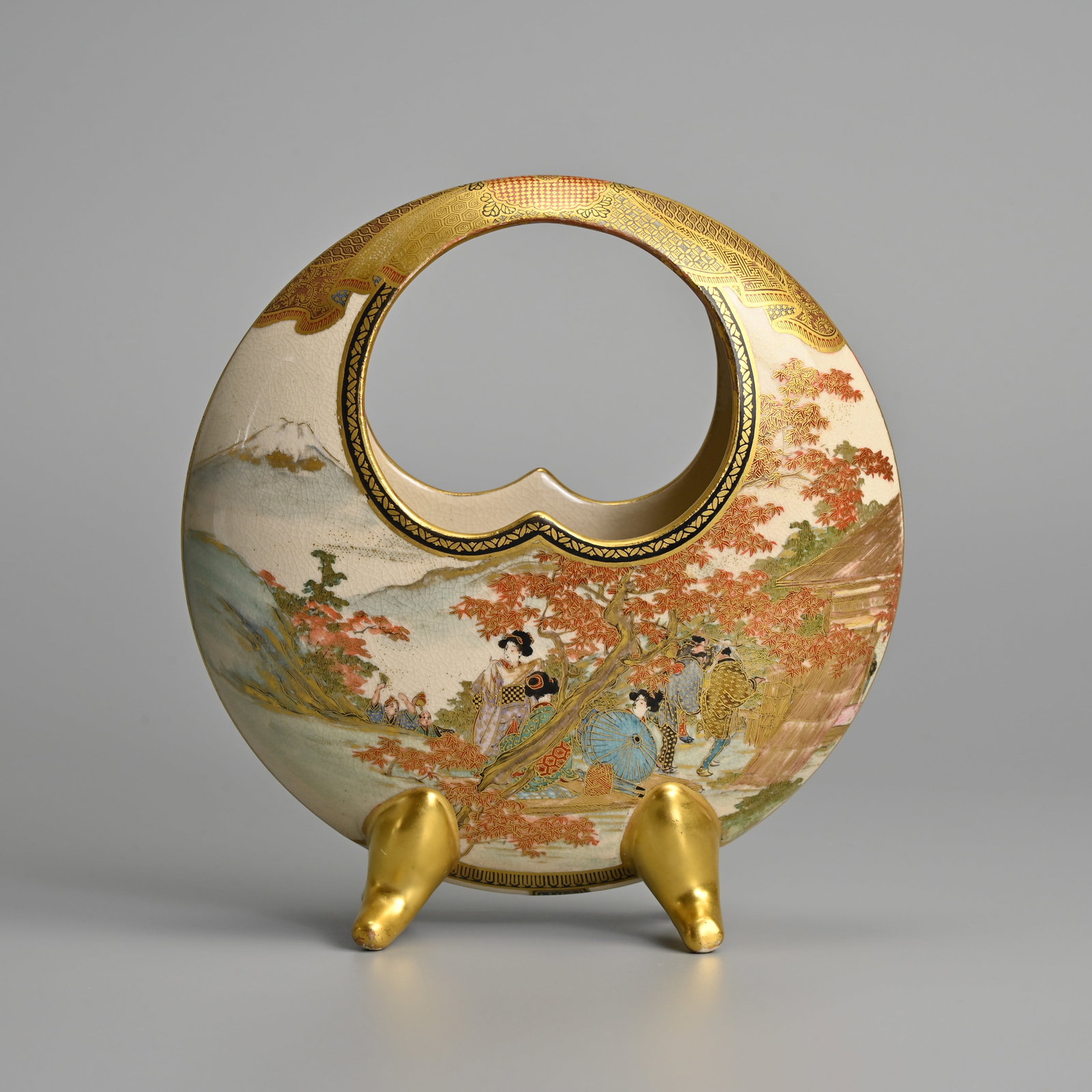 A JAPANESE SATSUMA WARE CIRCULAR HANDLED FOOTED VASE, IZUMIYAMA, LATE MEIJI PERIOD.: A JAPANESE SATSUMA WARE CIRCULAR HANDLED FOOTED VASE, IZUMIYAMA, LATE MEIJI PERIOD. Decorated with geisha behind trees and figures in landscape scene, the reverse with a rooster on a flowering cherry 