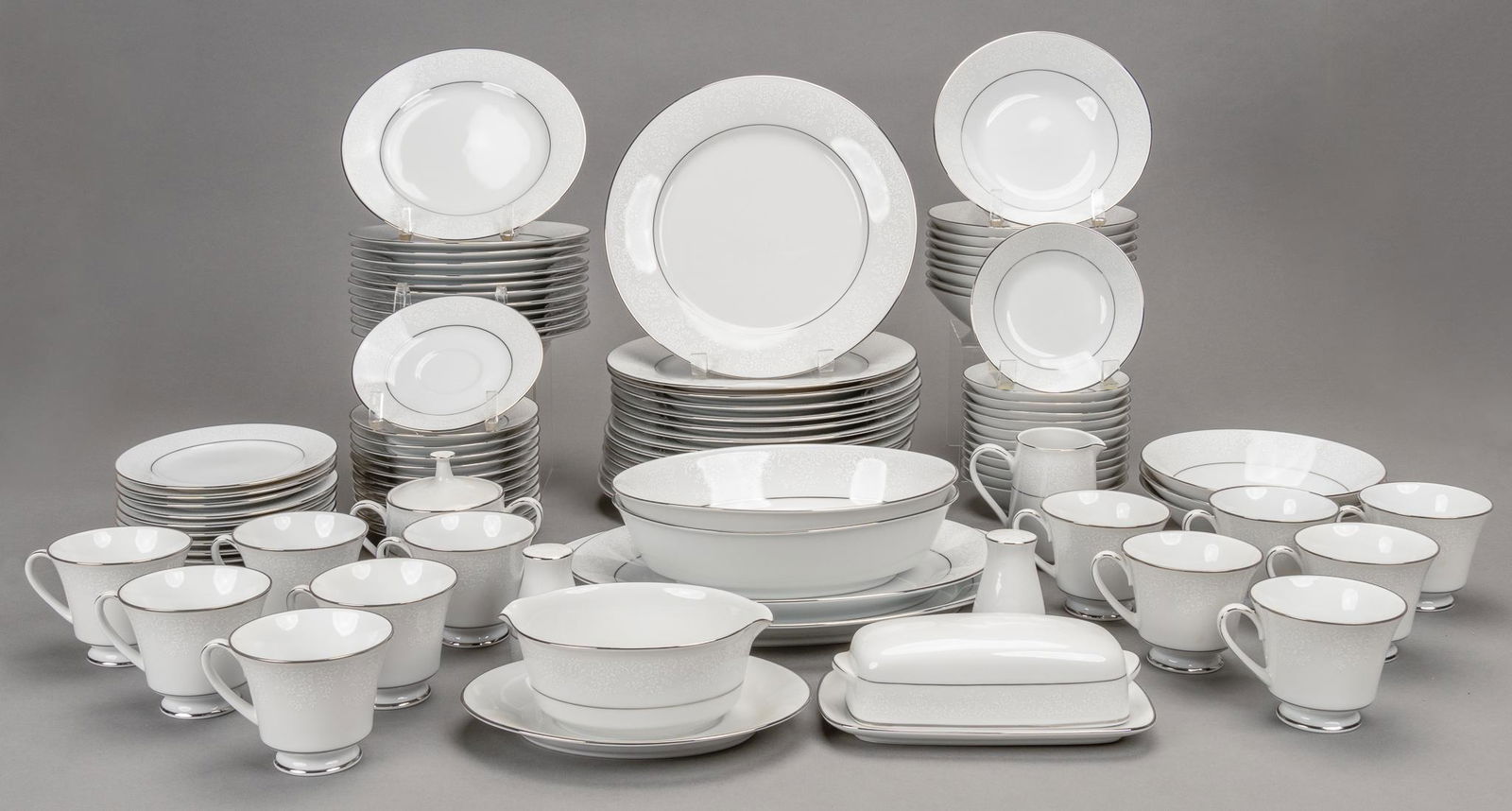 Noritake China Dinner Set: Large set of Japanese porcelain dinnerware, manufactured by Noritake, featuring floral motif on white body, metallic accents, to include teacups, butter dish, serving plates, chargers, serving dishes,