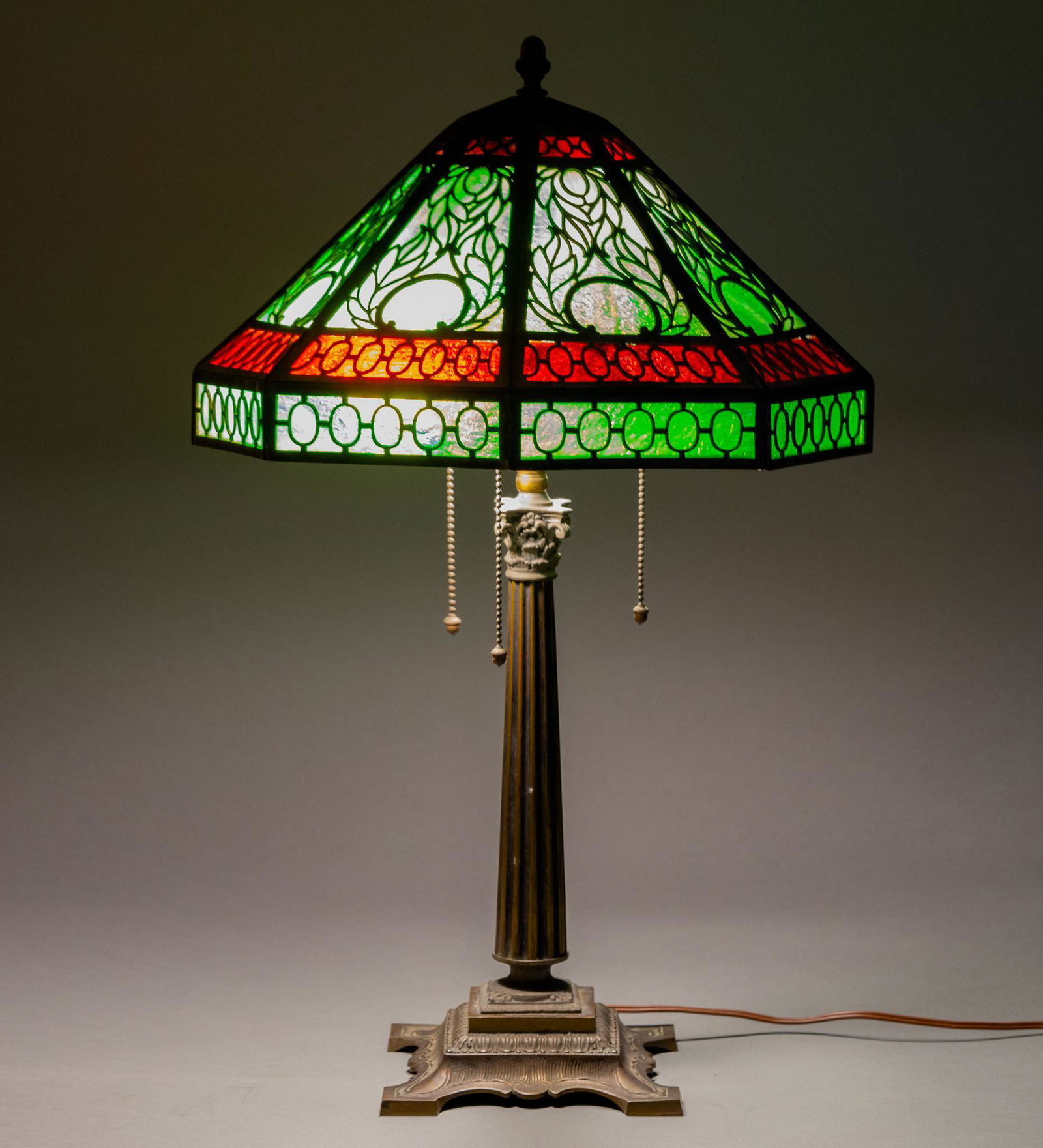 Arts and Crafts Panel Glass Lamp: Antique table lamp with two color panel glass shade on brass patinated iron base with columnar center and three light sockets with acorn pulls. Height 22 1/2 in., shade diameter at widest 14 in. Con