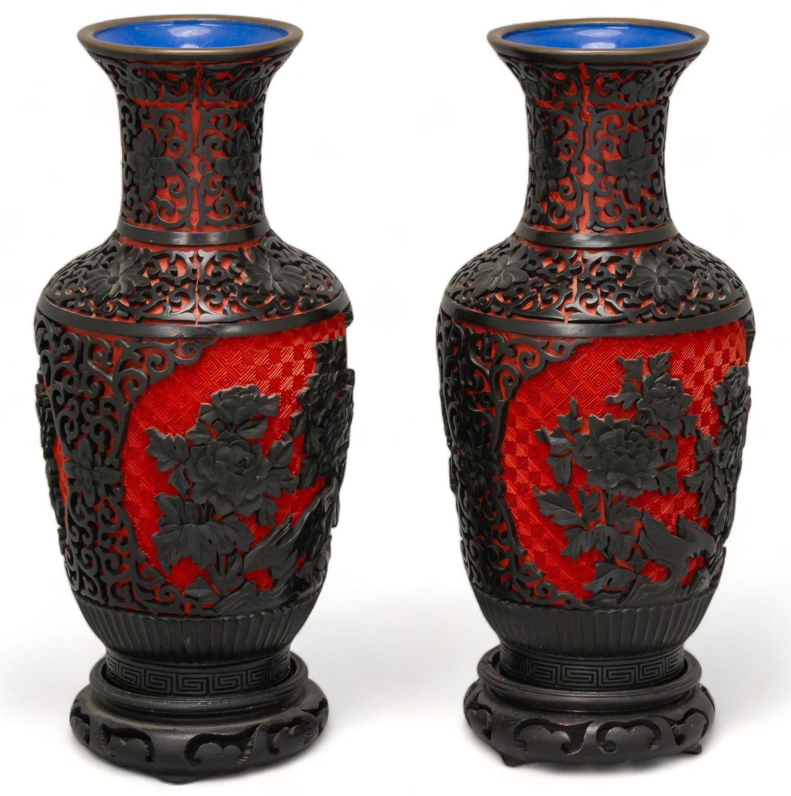 Vikki Carr | Pair of Chinese Cinnabar Vases: Matched pair of Chinese cinnabar vases, each with reserves on each side, central foliate carving surrounded by lotus and scrollwork carving, brass rim with blue enamel interior and base, raised on two