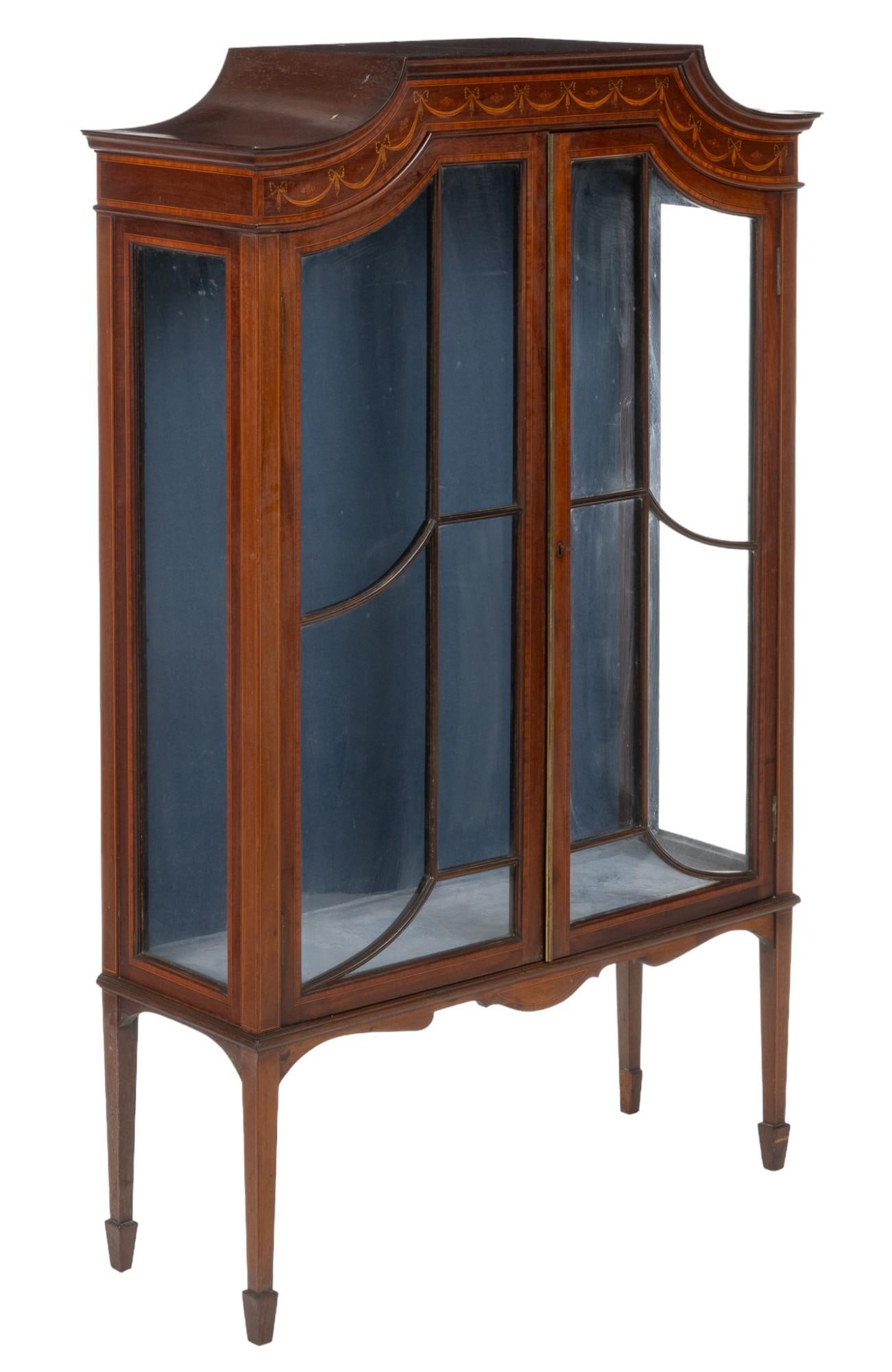 Regency Revival Inlaid Display Case: Regency revival inlaid display case featuring carved crest with painted ribbons and bows, geometric glass paneled doors opening to a blue velvet interior storage space with no shelves, on carved block