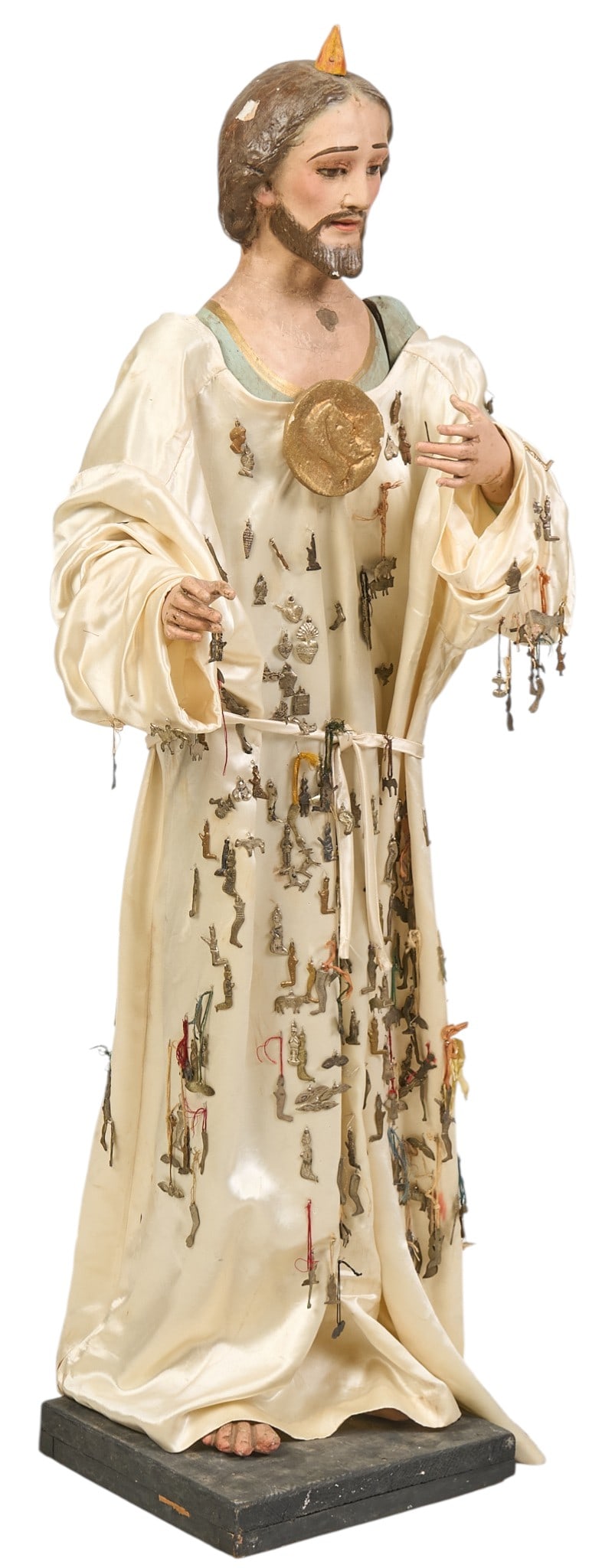 Jesus Christ Santo: Antique Spanish Colonial style polychrome Christ altar santo, hand-hand and painted, adorned with satin robe adorned with numerous milagro prayer charms, supported on stand, circa 19th to 20th century
