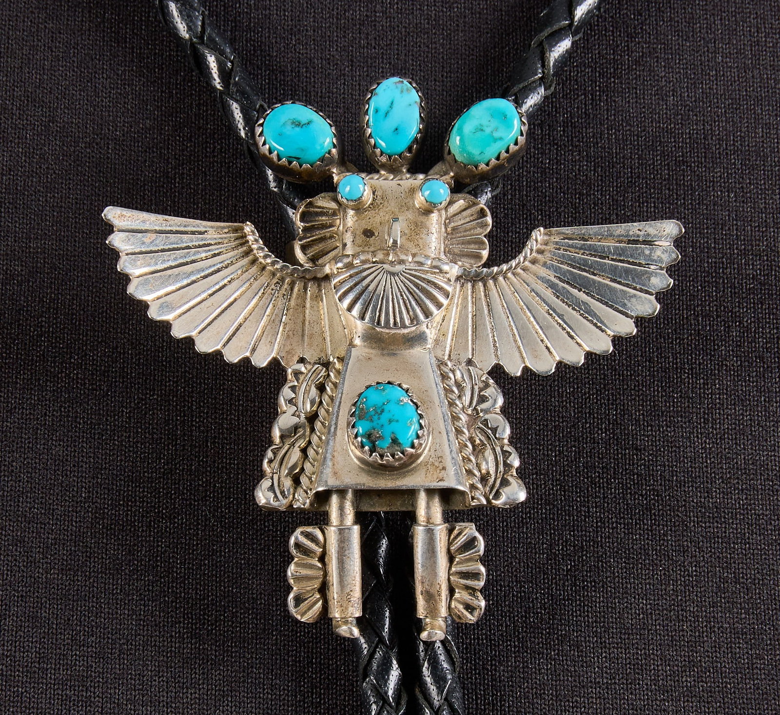 Navajo Sterling & Turquoise Kachina Bolo: Navajo sterling silver bolo tie, depicting kachina motif, set with turquoise cabochons, featuring twisted rope motifs and stamped details, on braided black leather cord, signed "RL" to rever