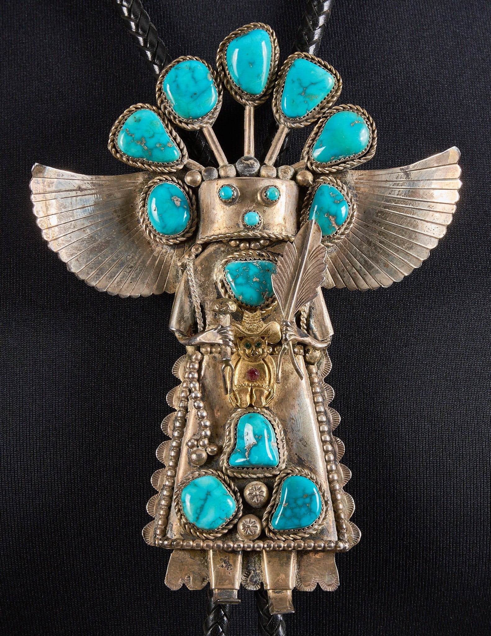 Sterling Silver & Turquoise Kachina Bolo Tie: Sterling silver bolo tie depicting kachina figure, set with turquoise stones and traditional Native American motifs, featuring gold-toned detail to center, set with green and red gemstones, on braided