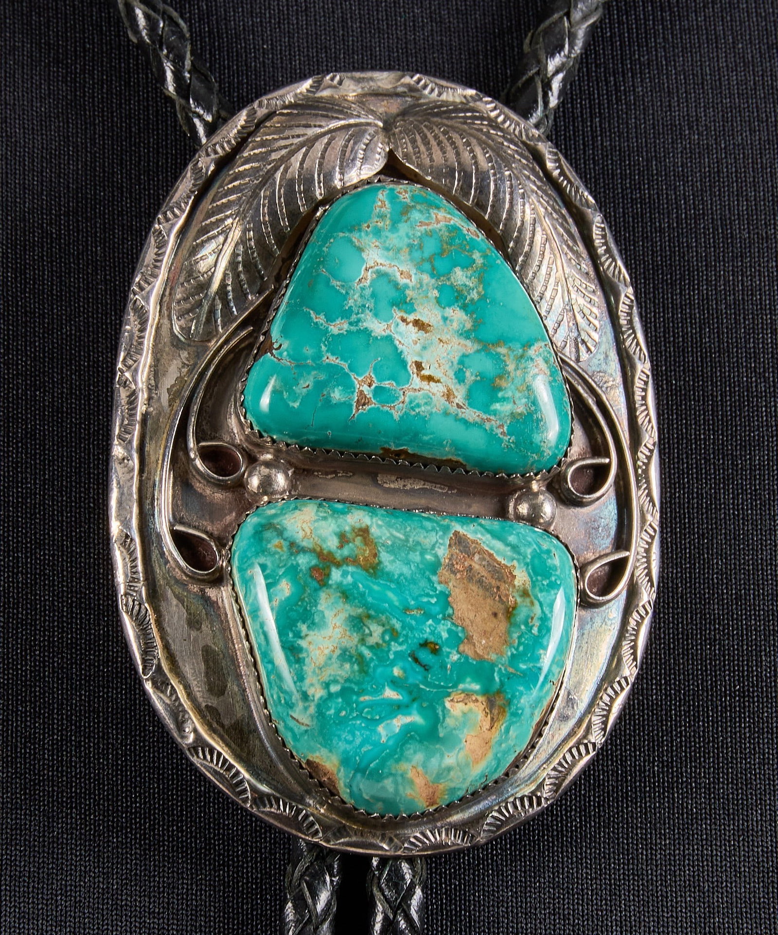 Southwestern Sterling & Turquoise Bolo Tie: Sterling silver bolo tie, set with two large turquoise stones, featuring feather motifs and stamped details, on braided black leather cord, signed Length: 39 in. Total weight: 72.8 g Provena