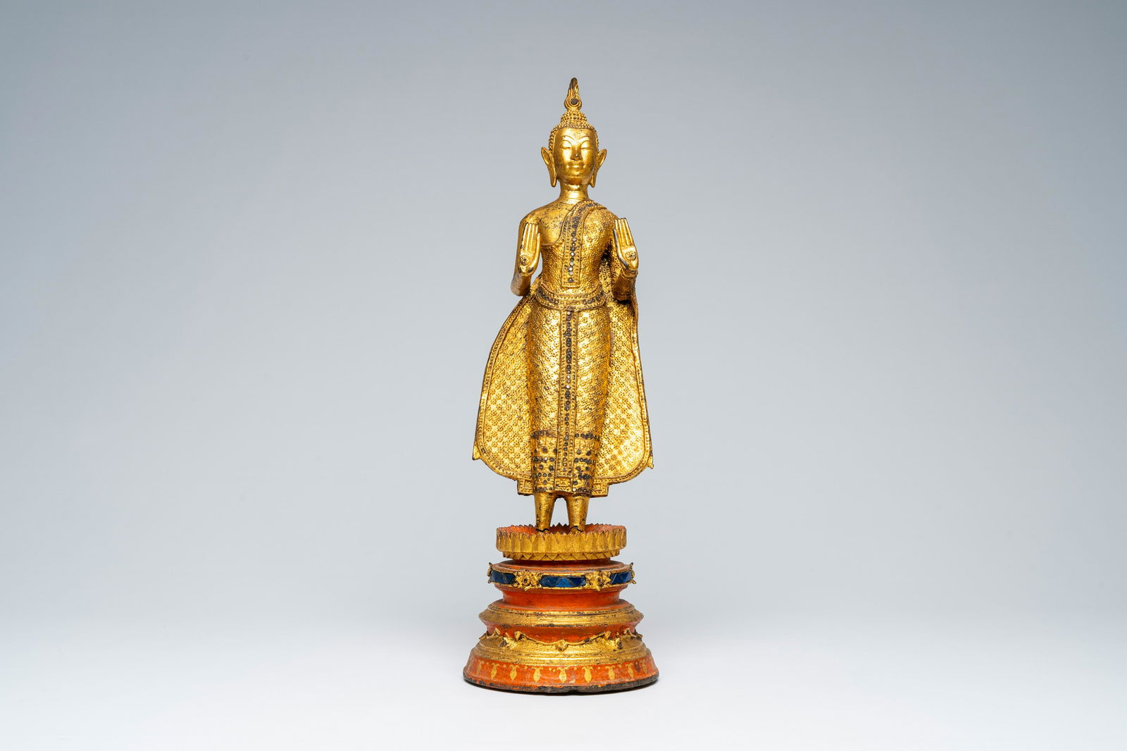 A Thai gilt bronze figure of a standing Buddha on a polychrome painted and glass inlaid metal base, (1 of 7)
