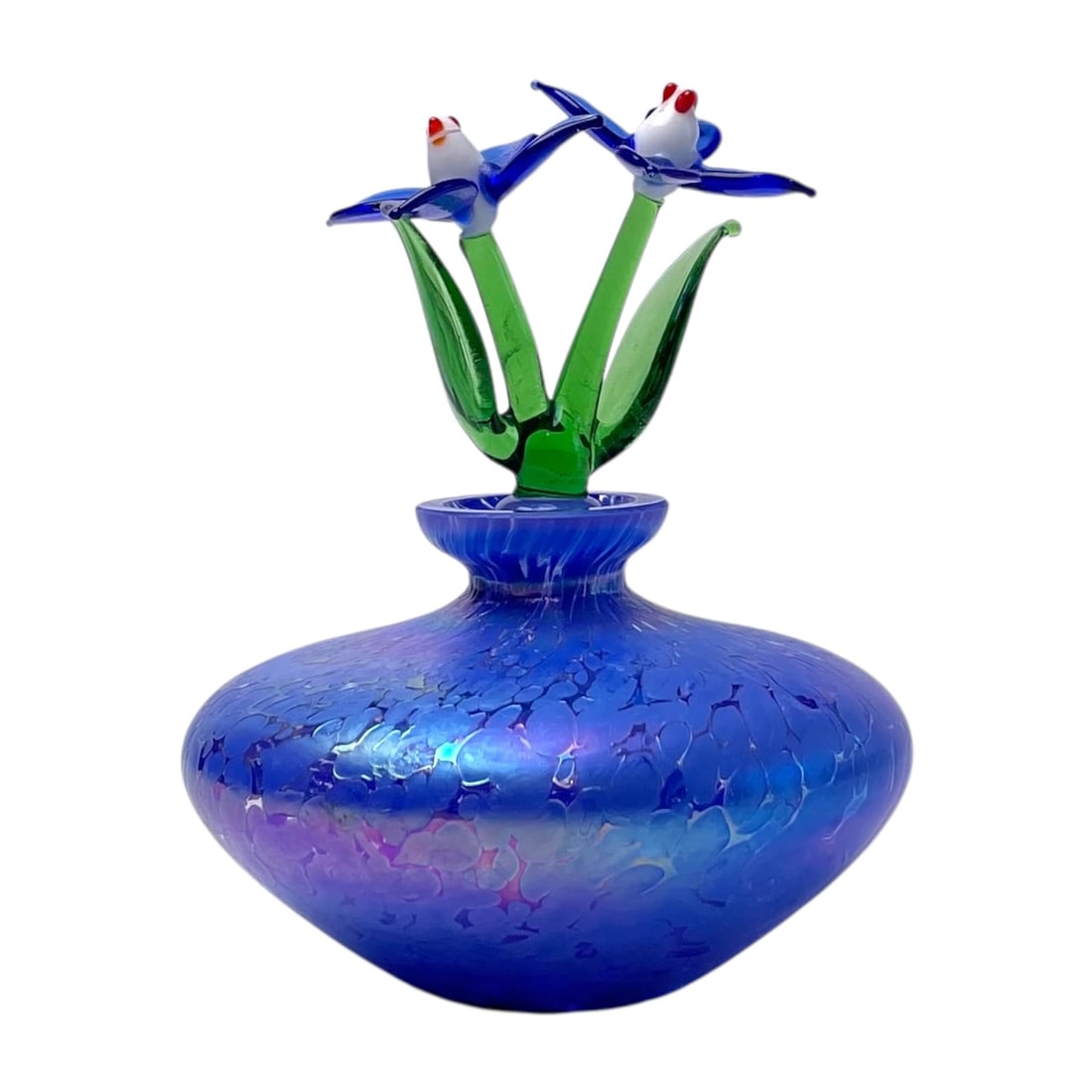 Caesars World Iridescent Art Glass Perfume Bottle: The bottle features a flattened, squat bulbous shape crafted from a blue iridescent flass. The top stopper is a detailed lampworked art glass shaped like green flower stems, blooming into blue petals 