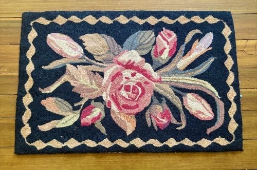 American Folk Art Floral Hooked Wool Rug 21"X32": A rectangular American folk art hooked rug featuring a central floral composition on a black field. The primary motif consists of a large, multi-petaled rose in variegated shades of crimson, pink, and
