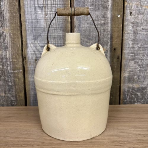 Antique Stoneware Jug with Wire Bail Handle and Wooden Grip: This antique stoneware jug features a cylindrical body with a rounded shoulder and a short, narrow neck. The exterior is finished in a monochromatic, cream-colored Bristol glaze. It is equipped with t