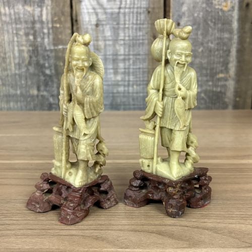 A Pair of Chinese Carved Soapstone Fisherman Figurines on Wooden Bases: A pair of hand-carved Chinese figurines crafted from pale olive-green soapstone. The sculptures depict joyful elderly fishermen, a traditional theme in Chinese decorative art representing abundance an