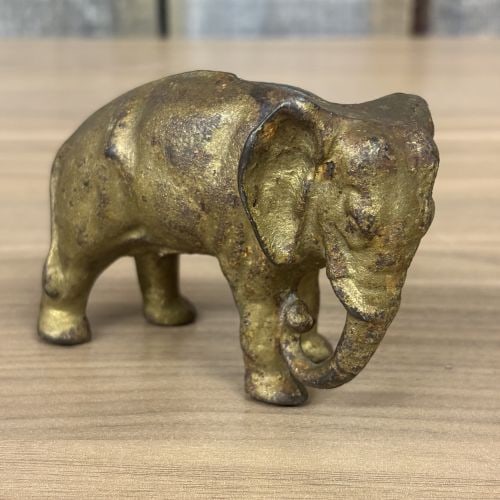 Cast Iron Elephant Still Bank in the Style of A.C. Williams: This antique still bank is constructed of cast iron and finished with a gold-tone bronzed wash. The piece is cast in the form of a standing elephant with its trunk lowered and curled inward. It featur