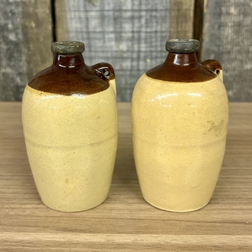 A Pair of Miniature USA Two-Tone Stoneware Whiskey Jug Salt and Pepper Shakers: This lot features a pair of matching miniature stoneware whiskey jugs designed for use as salt and pepper shakers. Each jug is formed with a bulbous body, a short neck, and a small applied strap handl