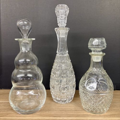 A Trio of Assorted Vintage Clear Glass Decanters: An assortment of three vintage clear glass decanters with matching stoppers. The collection includes a modernist-style decanter featuring a triple-lobed bubble-form body and a solid teardrop-shaped st