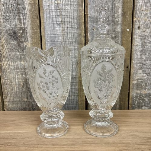A Pair of Mid-Century Pressed Colorless Glass Vessels in the Frosted Rose Pattern: A set of two matching mid-century pressed colorless glass vessels, comprising a tall lidded candy jar and a handled pitcher. Both pieces are decorated in the Frosted Rose pattern, which features high-