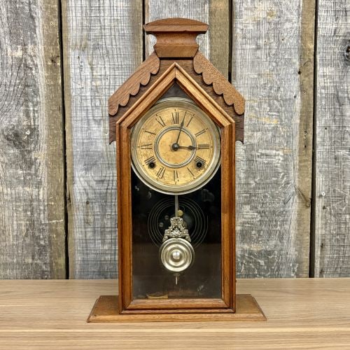 Antique Wood Chapel Style Mantel Clock Attributed to Waterbury Clock Co.: This late 19th-century mechanical shelf clock features a wooden case designed in the Gothic-influenced steeple or chapel style. The case is detailed with a scalloped pediment, a central pointed arch, 