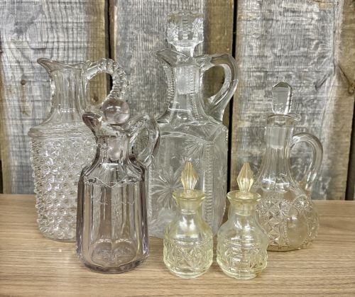 A Collection of Vintage American Pressed Glass Cruets and Miniature Bottles: A collection of vintage American pressed glass featuring various traditional patterns and forms. The assortment includes a pitcher with an all-over hobnail pattern and tiered handle, a large square-bo