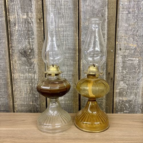 A Pair of Plume and Atwood Glass Kerosene Lamps with Dorset Burners: This pair of American pedestal-style kerosene lamps features pressed glass bodies and brass-toned metal burners. One lamp is made of clear glass, while the second lamp is composed of amber glass. Both