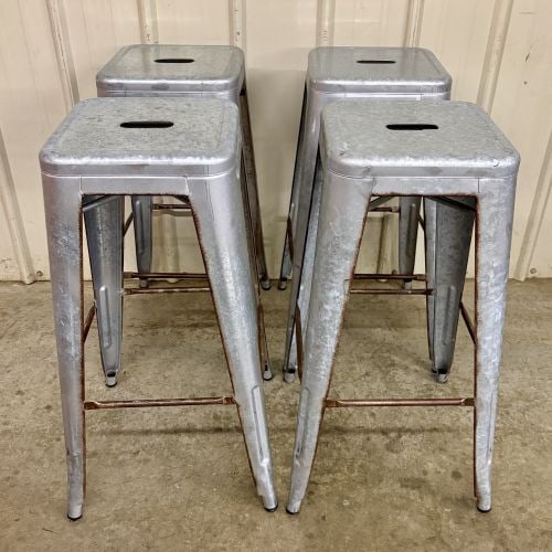 A Set of Four Industrial Distressed Metal Backless Bar Stools in the AmeriHome Loft Style: This set of four backless bar stools is designed in the industrial Marais or Tolix style, closely resembling the AmeriHome Loft series. The stools are constructed from metal featuring a silver-toned g