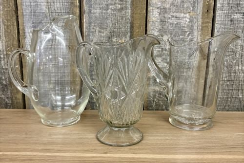 A Trio of Vintage Clear Glass Water Pitchers: This lot comprises three clear glass water pitchers of varying styles, dating from the mid-20th century. The first item is a large ball-shaped pitcher featuring a bulbous body, a hand-applied handle w