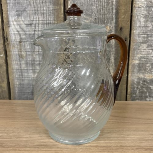 Vintage Depression Era Spiral Optic Glass Pitcher with Applied Amber Handle and Matching Lid: This lidded glass pitcher is constructed of clear glass featuring a molded diagonal spiral optic pattern throughout the body and lid. The piece is accented with an applied amber glass handle and a mat