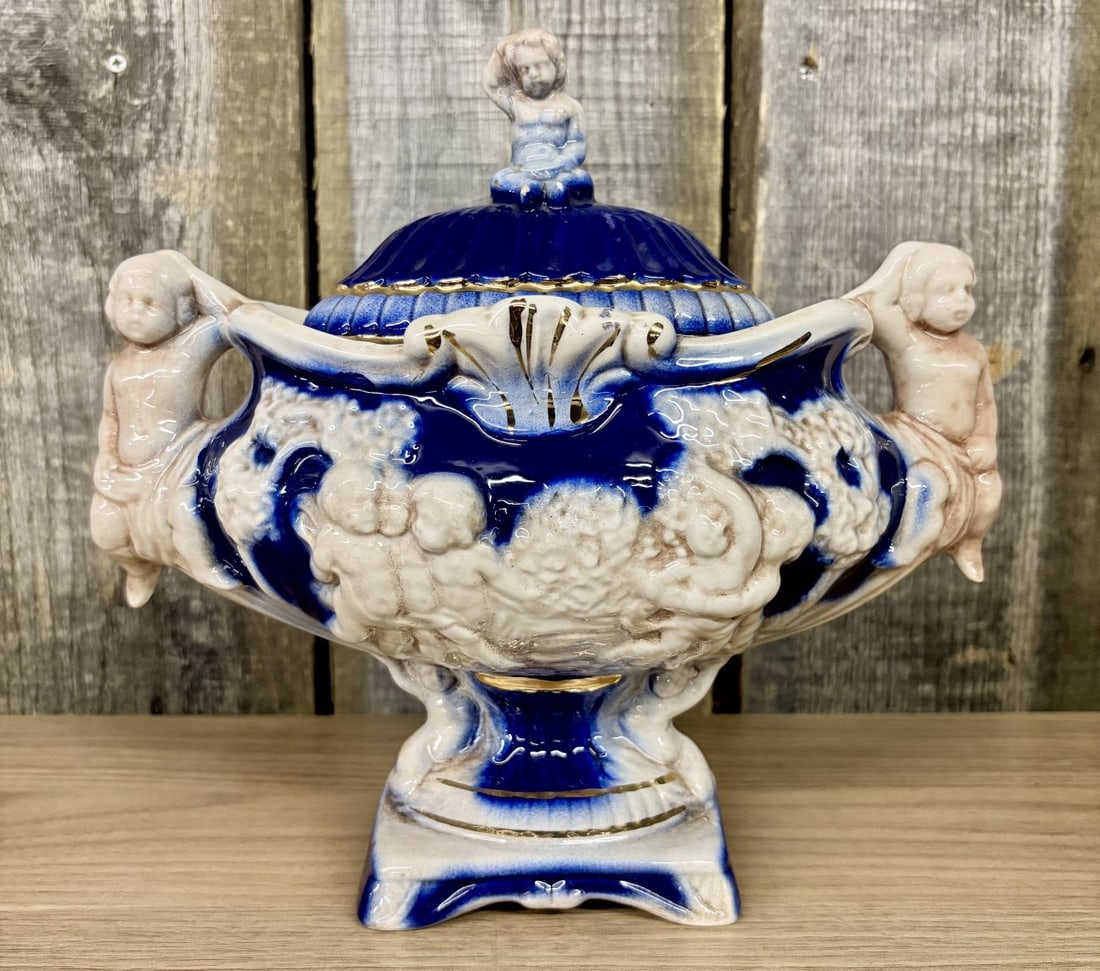Norleans Italian Ceramic Cobalt Blue and Gold Cherub Lidded Urn: This is a footed and lidded ceramic decorative urn produced in Italy and distributed by Norleans. The vessel features a boat-shaped body supported by a pedestal base. It is decorated with high-relief 