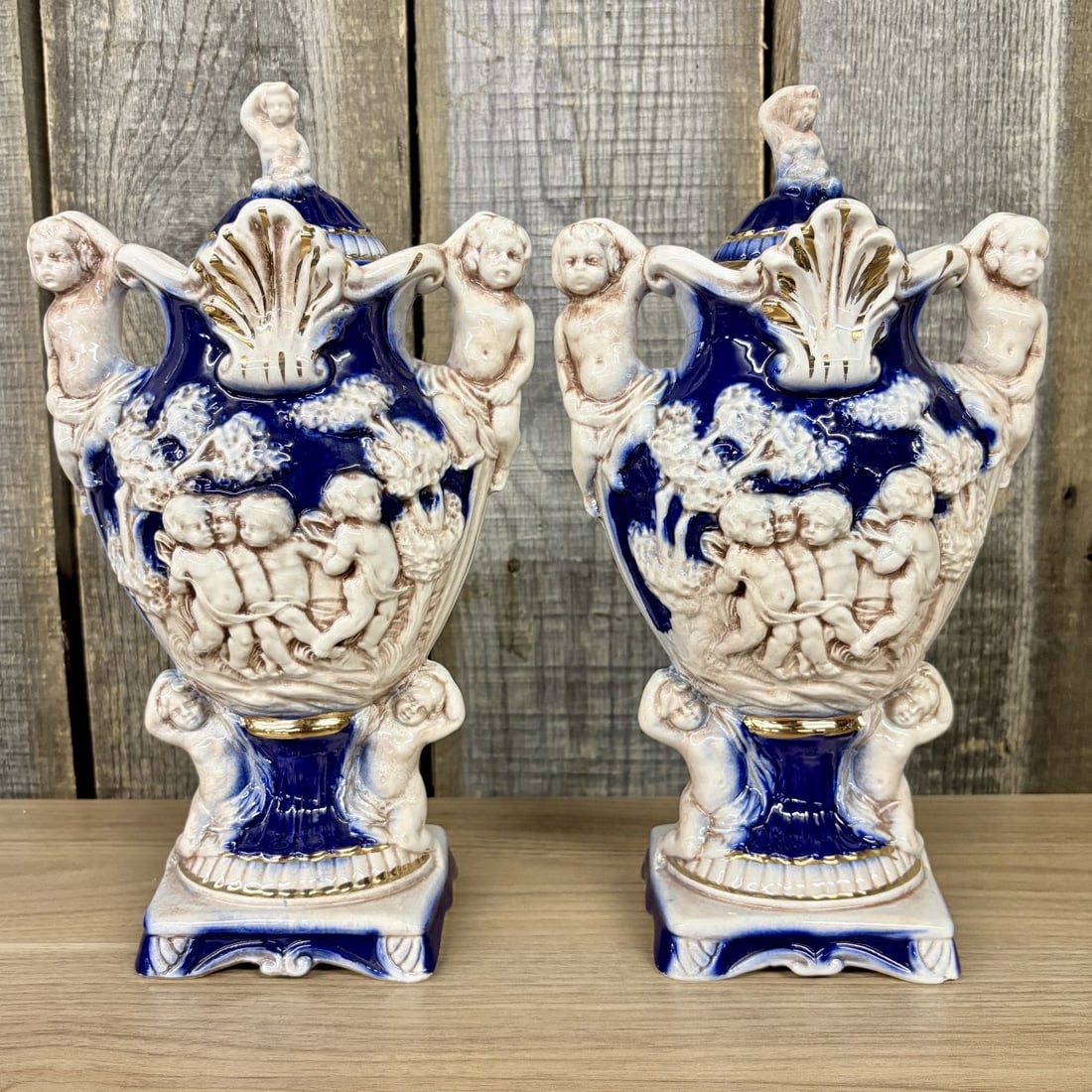 A Pair of Norleans Italy Cobalt Blue and Gilt Ceramic Lidded Urns with Cherub Relief: A pair of decorative ceramic lidded urns manufactured in Italy and imported under the Norleans brand. Each vessel is of baluster form, resting on a square pedestal base. The urns feature extensive hig