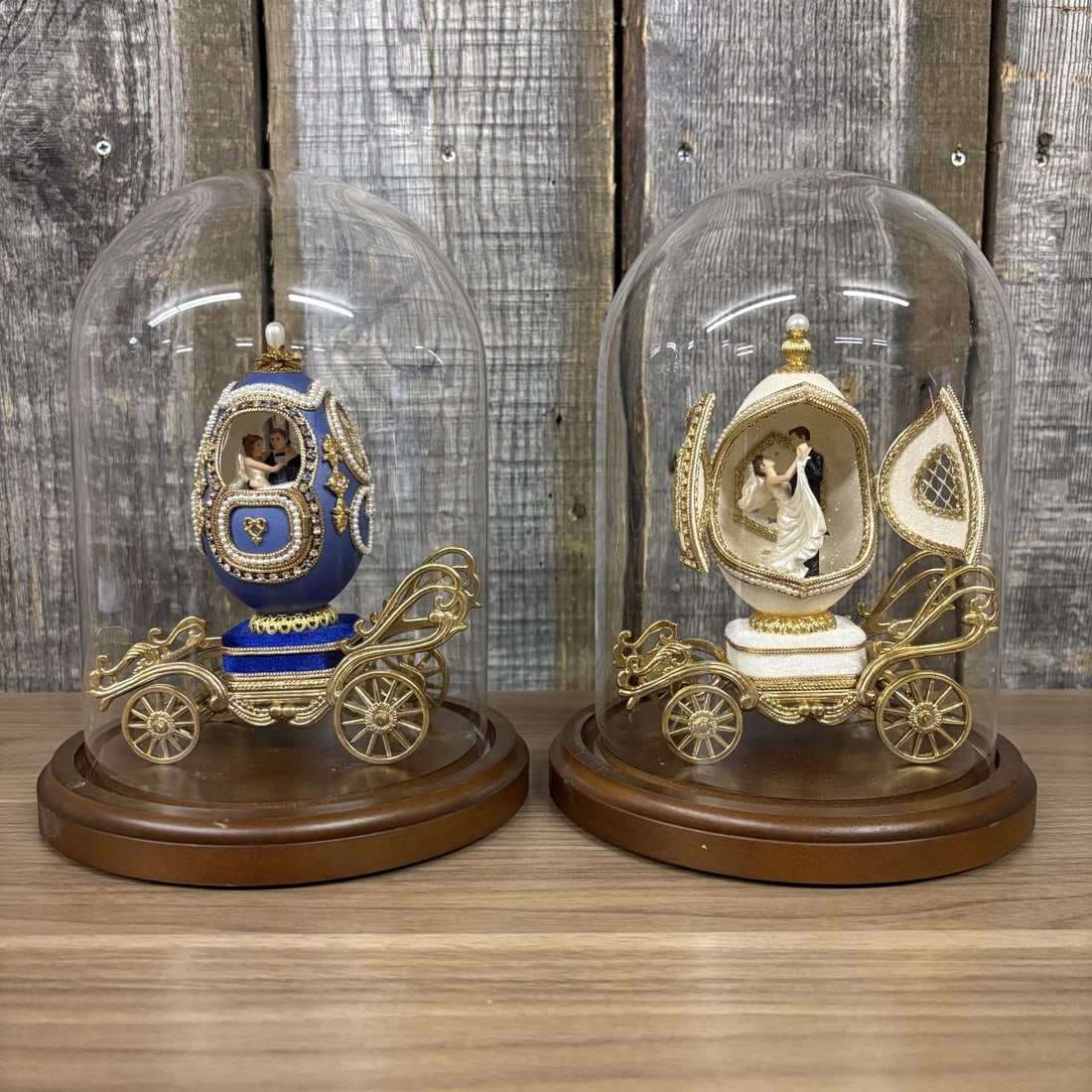 A Pair of Musical Faberge Style Wedding Carriage Egg Figurines with Glass Cloches: This lot consists of two ornate wedding-themed musical egg carriages, each presented under a glass cloche on a circular wooden base. These pieces are designed in the tradition of Faberge-style egg art