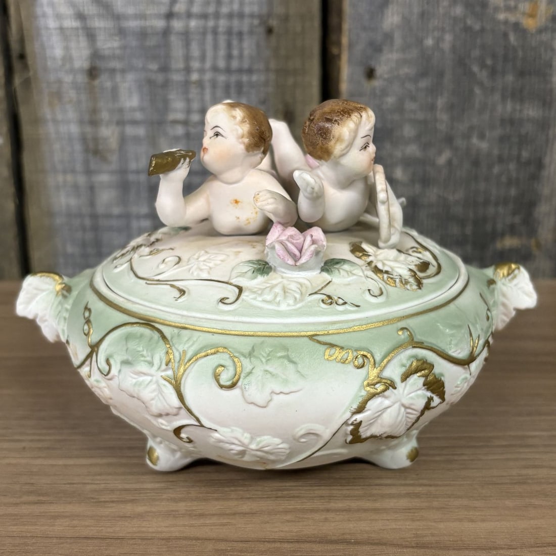 Norleans Japan Porcelain Cherub Figural Trinket Box: This porcelain lidded trinket box was imported by Norleans. The vessel features an oval, footed form with twin leaf-style handles. The domed lid is surmounted by two bisque-finished cherub figures, on