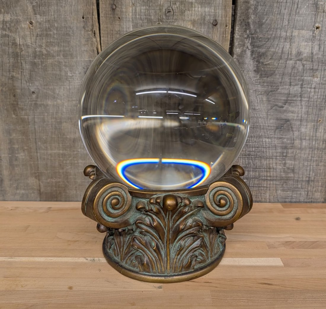 Large Optical Glass Sphere on Neoclassical Patinated Metal Base: This decorative object features a substantial solid clear glass sphere resting on an ornate neoclassical style base. The sphere is crafted from high-quality optical glass, polished to a reflective sur