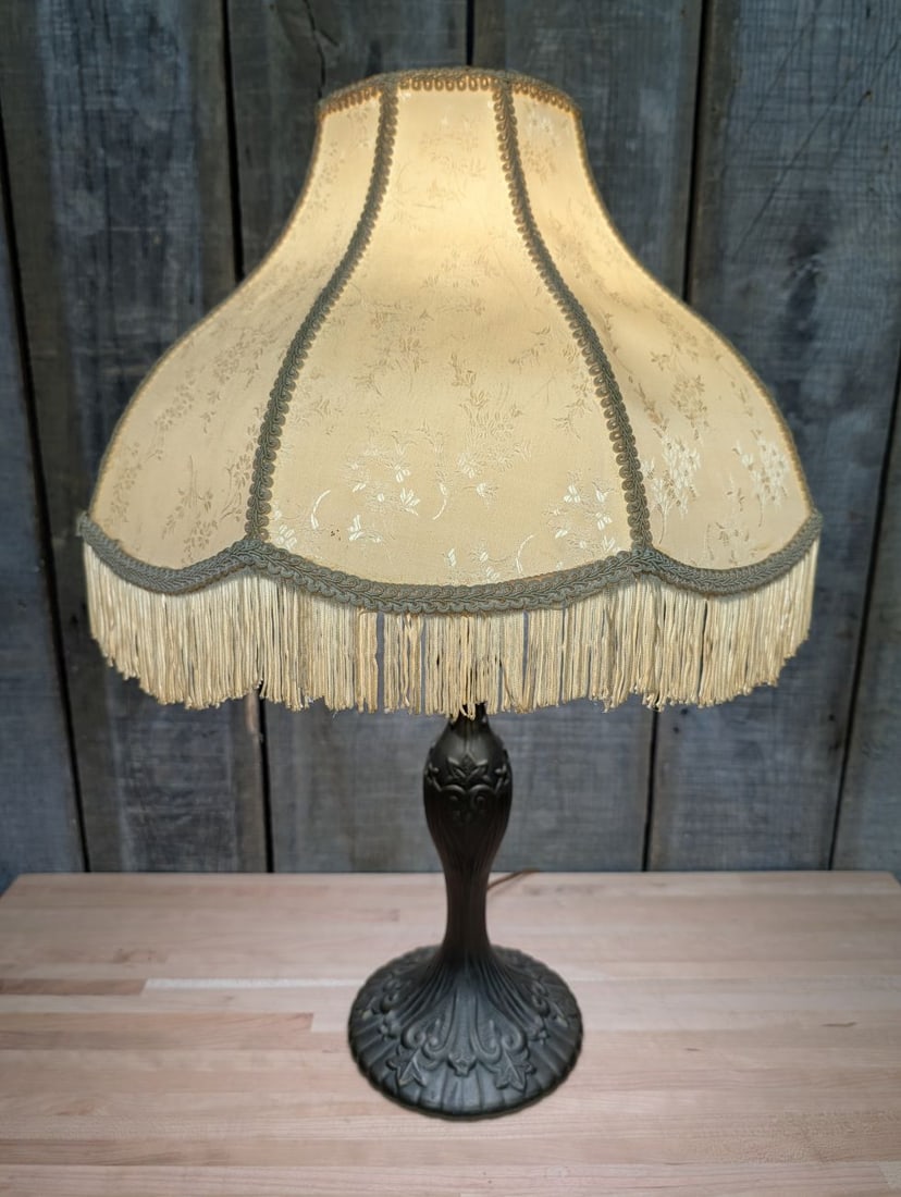 Victorian Style Cast Metal Table Lamp with Fringed Fabric Shade: This table lamp features a decorative cast metal base and stem finished with a dark bronze-toned patina. The circular base has a scalloped, lobed edge and is ornamented with raised foliate and scrollw