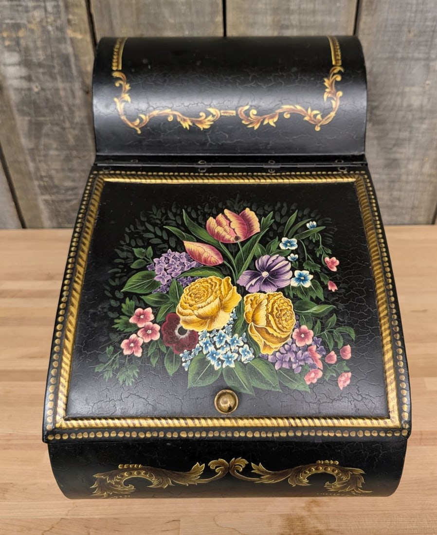 Victorian Style Hand Painted Toleware Floral Coal Bin: This fireside coal bin is constructed from fabricated sheet metal with a black japanned finish. The piece features a slanted, hinged lid and an arched top section. The lid is decorated with a central 