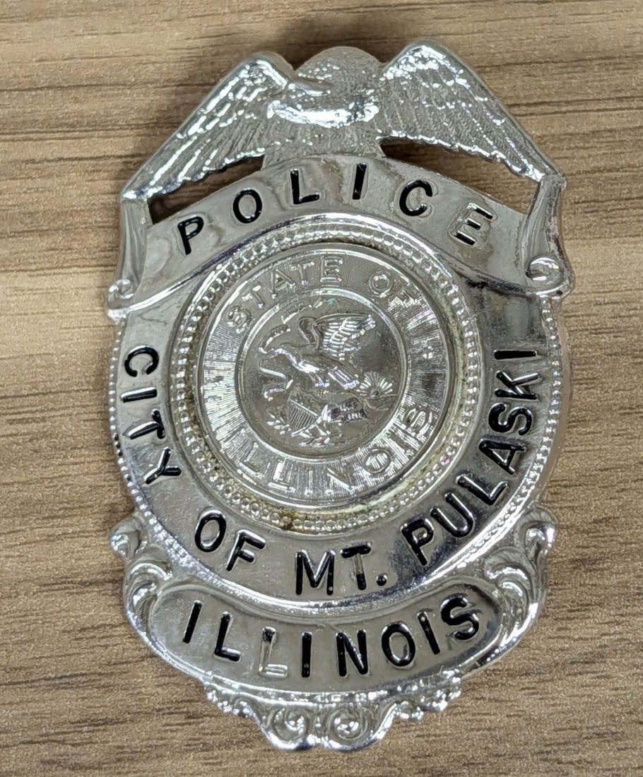 City of Mt. Pulaski Illinois Police Department Eagle Top Shield Badge: A shield-form law enforcement badge representing the City of Mt. Pulaski, Illinois. The badge is surmounted by a spread-winged eagle crest. The central circular medallion displays the Great Seal of th