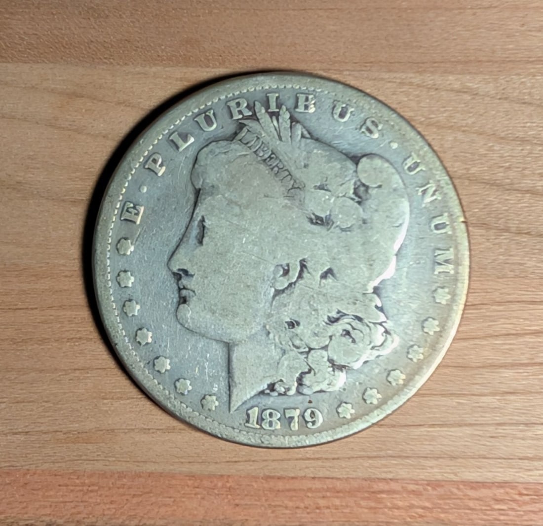 1879 US Morgan Silver Dollar: This US 1879 Morgan silver dollar was struck at the Philadelphia mint. The coin is composed of 90% silver and 10% copper. The obverse features the Liberty head design, while the reverse displays the h