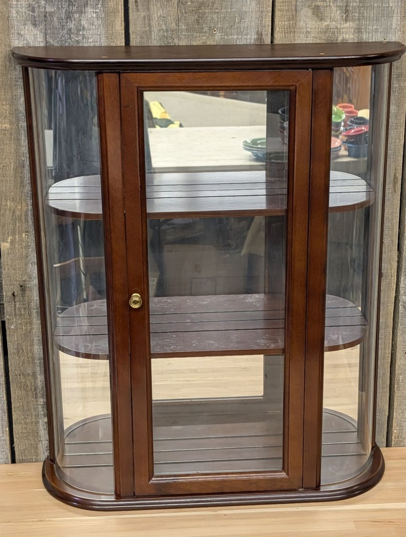 The Bombay Company Mahogany Tone Bow Front Wall Curio Cabinet with Decorative Brackets: A one piece wall mounted curio display cabinet manufactured by The Bombay Company. The cabinet features a bow front design with curved glass side panels and a central glass door within a wood frame. T