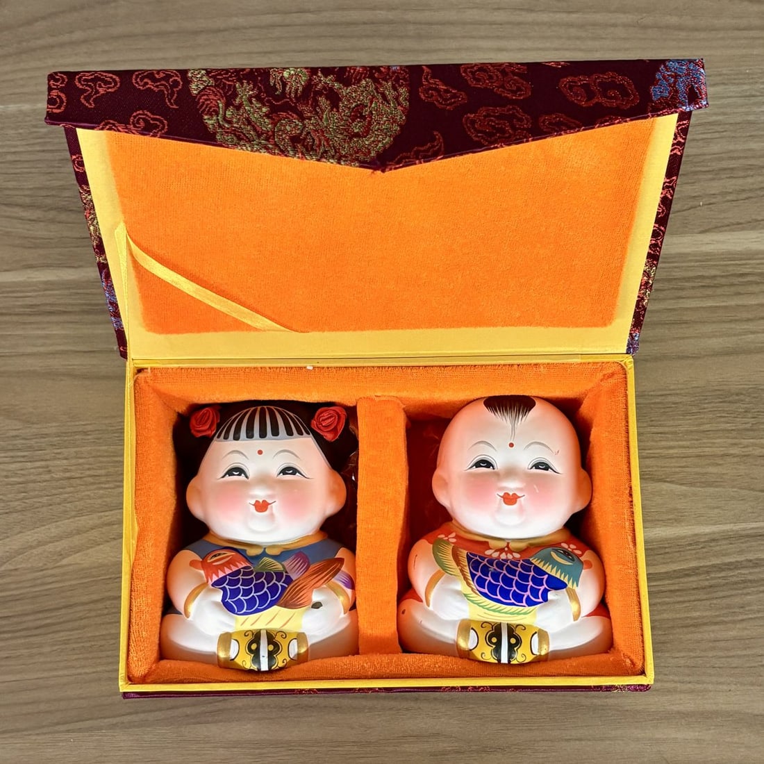 A Pair of Wuxi Huishan Da A Fu Clay Figurines in Presentation Box: A pair of Huishan clay figurines known as Da A Fu (Great Fortune), originating from Wuxi, Jiangsu Province, China. The set comprises a boy and a girl figurine, both depicted in a seated position holdi