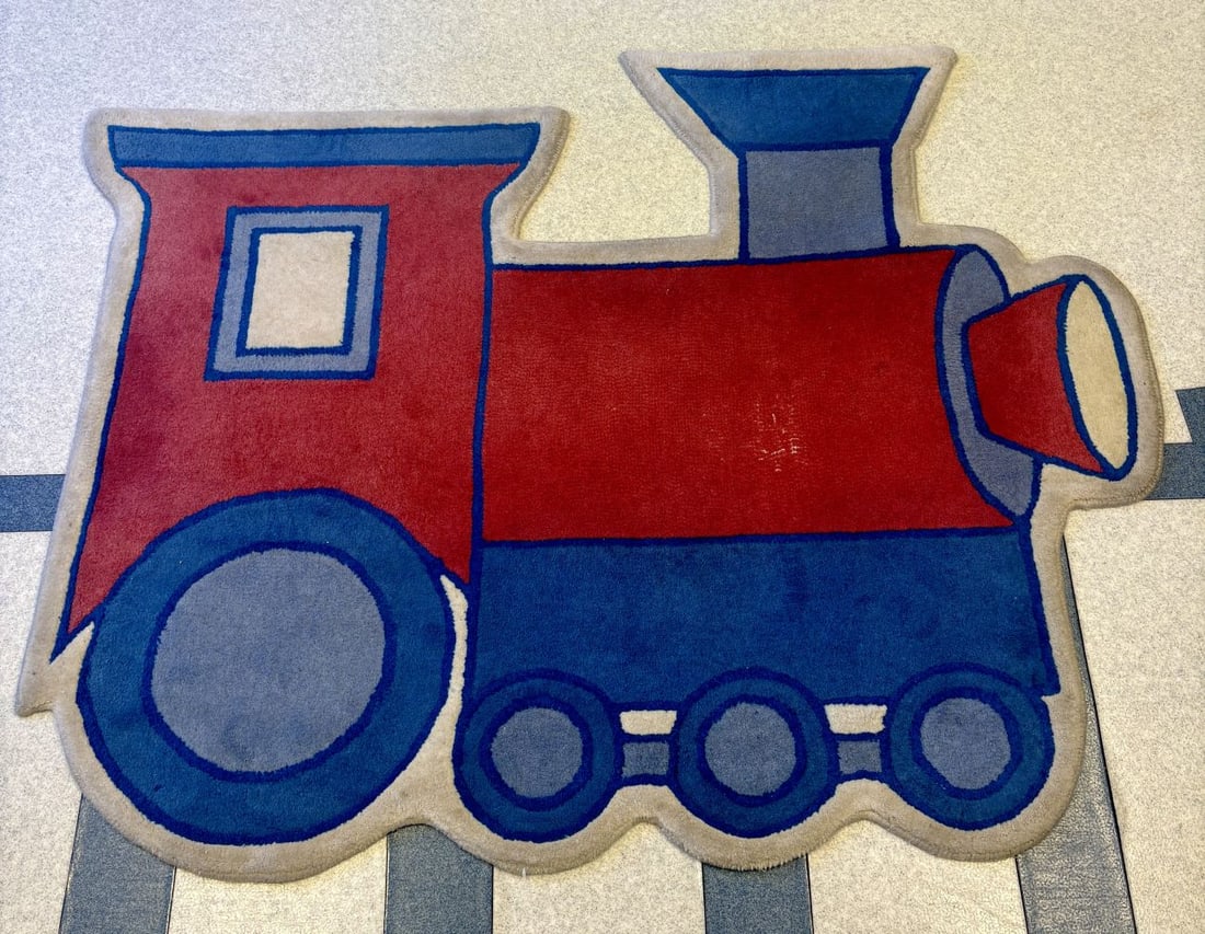 A Novelty Shaped Steam Locomotive Play Rug: A large novelty shaped rug depicting a steam locomotive engine in a vibrant primary color palette of red, blue, and cream. The design follows a traditional locomotive silhouette, featuring a large red