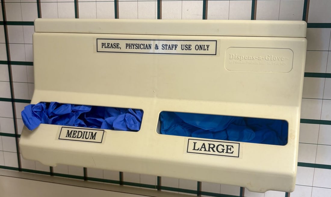 Pinnacle Products Inc. Dispens-a-Glove Wall-Mounted Medical Glove Dispenser: A wall-mounted bulk medical glove dispenser manufactured from molded cream-colored plastic. The unit features two front-facing dispensing slots for different sizes. An embossed rectangular panel on th
